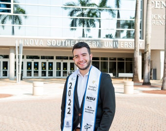 May include: A graduate in a black suit and white shirt stands in front of a university building. He wears a white and blue sash with the text "2022", "NSU Florida", and "B.S. Criminal Justice". The university name is visible in the background.