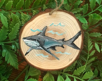 May include: A hand-painted wooden ornament depicting a grey shark swimming in blue water. The ornament is round and has a rustic wood grain texture. It is ideal for a nautical themed Christmas tree.
