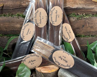 May include: Various incense sticks in clear packaging, branded as "Mystical Outpost" with scent labels such as "Rosemary Mint" and "All Spice." The incense is presented on a bed of pebbles and wood, with some greenery.