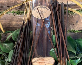 May include: Incense sticks in a clear plastic bag with a brown label that reads "Mystical Outpost". The incense sticks are dark brown and black, with a wooden background and small stones at the base. A small wooden stump is in the center.