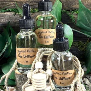 May include: Three clear glass car diffuser bottles with black droppers, labeled "Mystical OutPost Car Diffuser 1 oz." A wooden car air freshener hangs in front. The background includes greenery and a wooden surface.