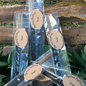 May include: Several packages of incense sticks are displayed against a natural wood and greenery backdrop. Each clear plastic package contains a bundle of dark incense sticks and is labeled with an oval tag that reads "Mystical Outpost Sample".