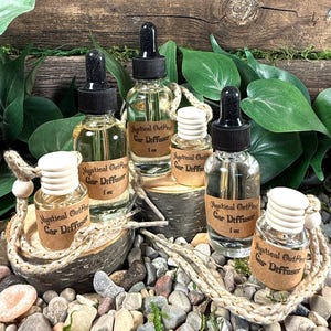 May include: Four glass bottles with droppers filled with clear liquid and labeled "Mystical OutPest Car Diffuser 1 oz" are displayed on a wooden surface with a background of green foliage and small stones.