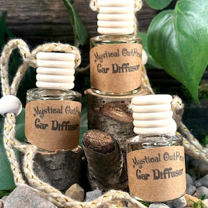 May include: Three glass bottles with brown paper labels that say "Mystical Outpost Car Diffuser". The bottles are hanging from a white string with a wooden bead at the end. The bottles are sitting on a wooden surface with rocks and green plants in the background.