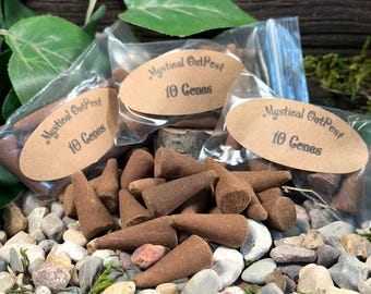 Pode incluir: Um close-up de um monte de cones de incenso marrons com três pequenas bolsas de cones de incenso. Cada bolsa tem um rótulo de papel castanho que diz "Mystical OutPest 10 Cones".
