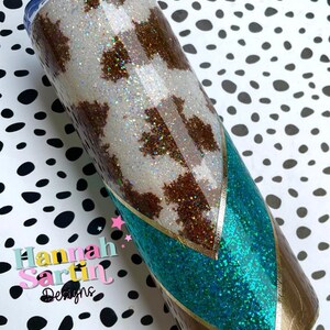 May include: A white and brown cow print tumbler with a turquoise and gold glitter design. The tumbler has a clear lid and a stainless steel body. The tumbler is on a white and black polka dot background.