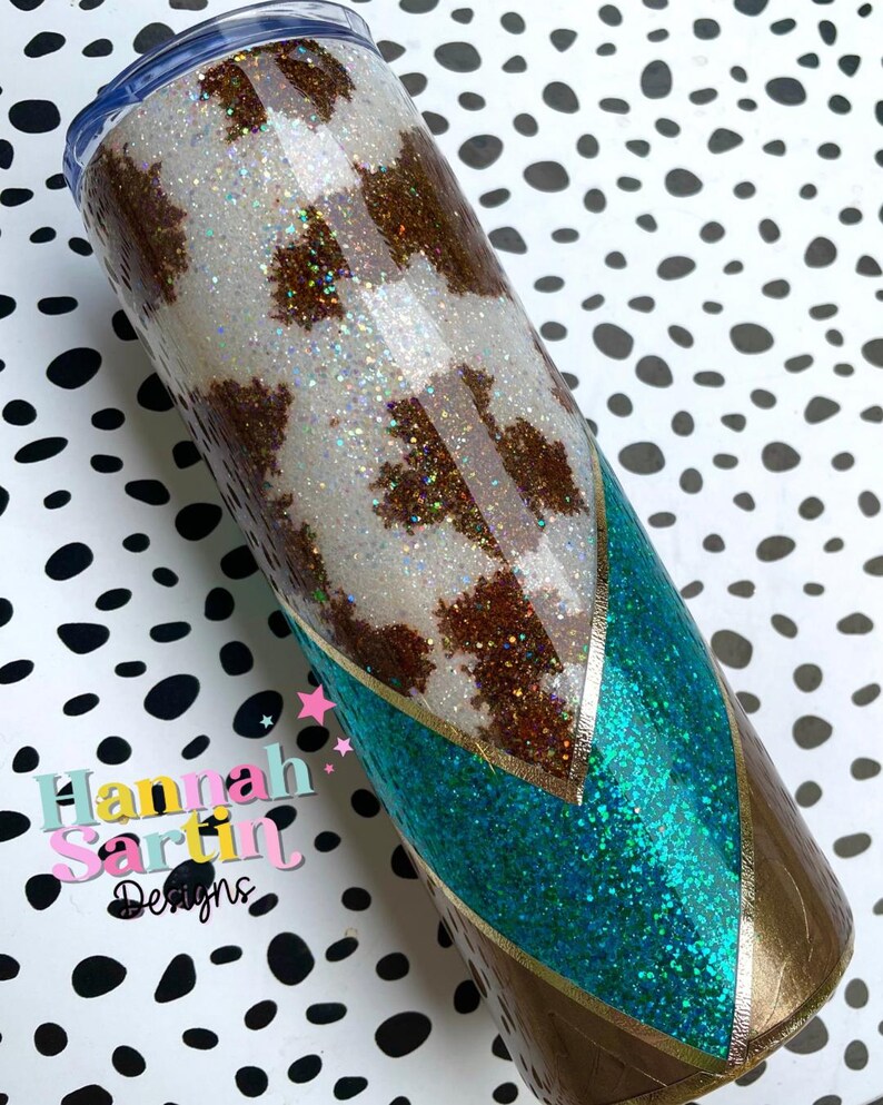 May include: A white and brown cow print tumbler with a turquoise and gold glitter design. The tumbler has a clear lid and a stainless steel body. The tumbler is on a white and black polka dot background.