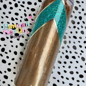 May include: A gold and turquoise glitter tumbler with a gold and white patterned design. The tumbler has a clear lid and a gold rim.