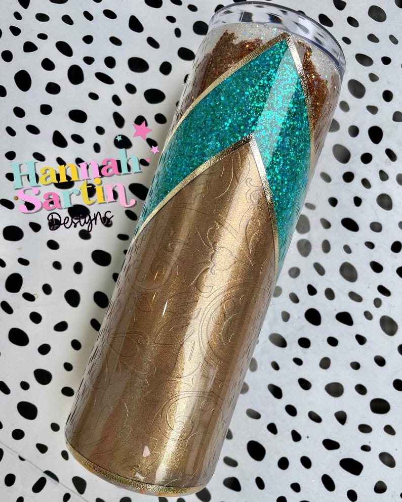 May include: A gold and turquoise glitter tumbler with a gold and white patterned design. The tumbler has a clear lid and a gold rim.