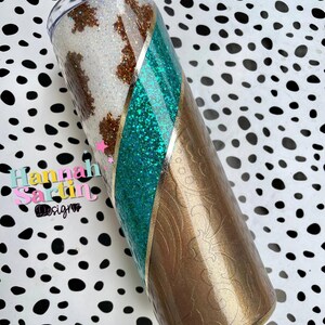 May include: A white and brown cow print tumbler with a teal glitter stripe and gold accents. The tumbler has a brown floral pattern on the bottom half.