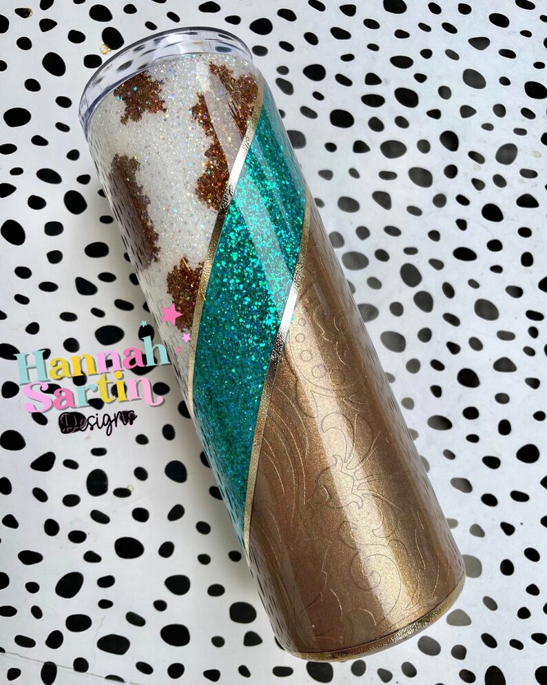 May include: A white and brown cow print tumbler with a teal glitter stripe and gold accents. The tumbler has a brown floral pattern on the bottom half.