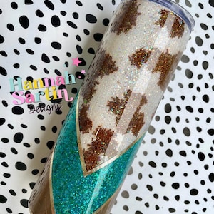 May include: A white and brown cow print tumbler with a turquoise and gold glitter design. The tumbler has a clear lid and is decorated with glitter.