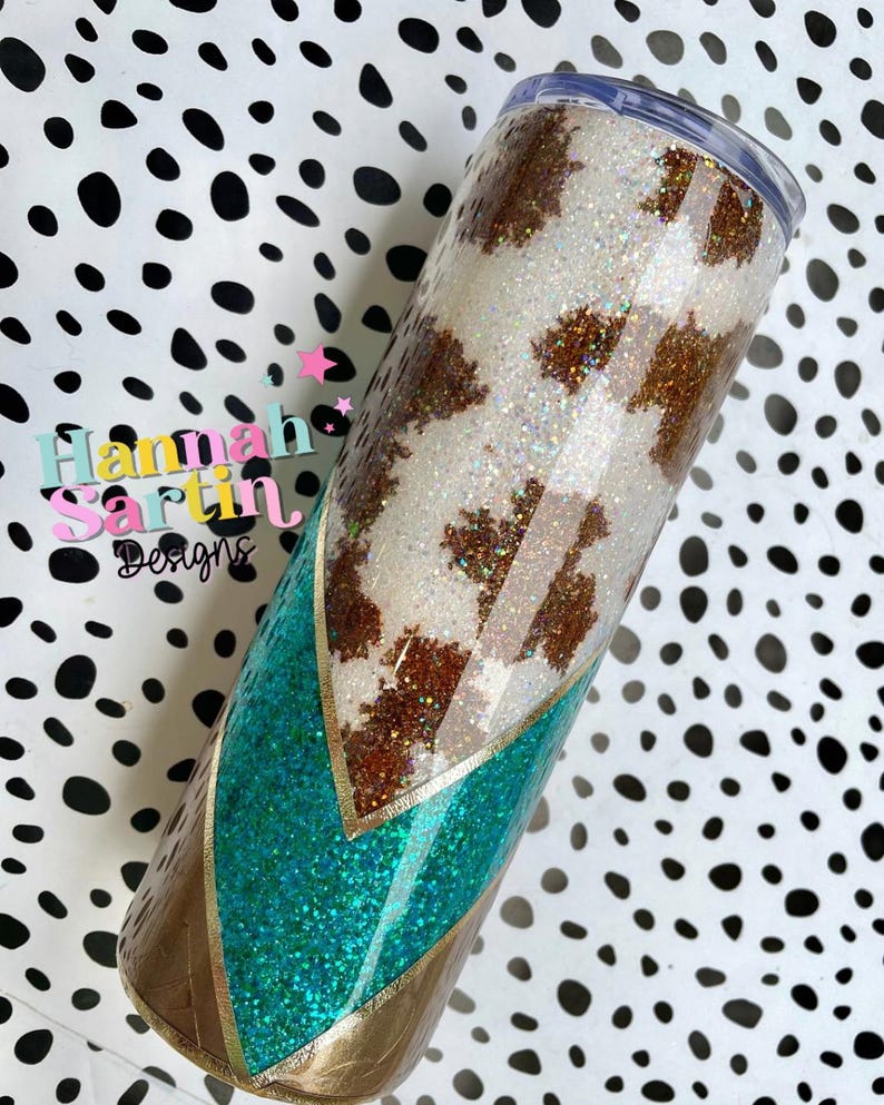 May include: A white and brown cow print tumbler with a turquoise and gold glitter design. The tumbler has a clear lid and is decorated with glitter.