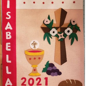 May include: A pink felt banner with the name "ISABELLA" in white letters on a red felt strip. The banner also features a brown felt cross with white flowers, a chalice, grapes, a loaf of bread, and the year "2021" in red felt. The banner is decorated with red and white felt dots around the edges.