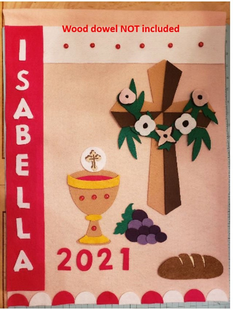 May include: A pink felt banner with the name "ISABELLA" in white letters on a red felt strip. The banner also features a brown felt cross with white flowers, a chalice, grapes, a loaf of bread, and the year "2021" in red felt. The banner is decorated with red and white felt dots around the edges.