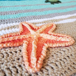 Peut inclure: Une &eacute;toile de mer en crochet dans des tons d'orange et de rose, avec une surface textur&eacute;e, est plac&eacute;e sur une couverture en crochet beige avec des rayures blanches, bleu clair et grises.