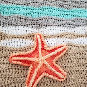 Peut inclure: Une couverture au crochet avec un motif ray&eacute; dans les tons de gris, blanc et turquoise. Une grande &eacute;toile de mer orange est appliqu&eacute;e sur la couverture.