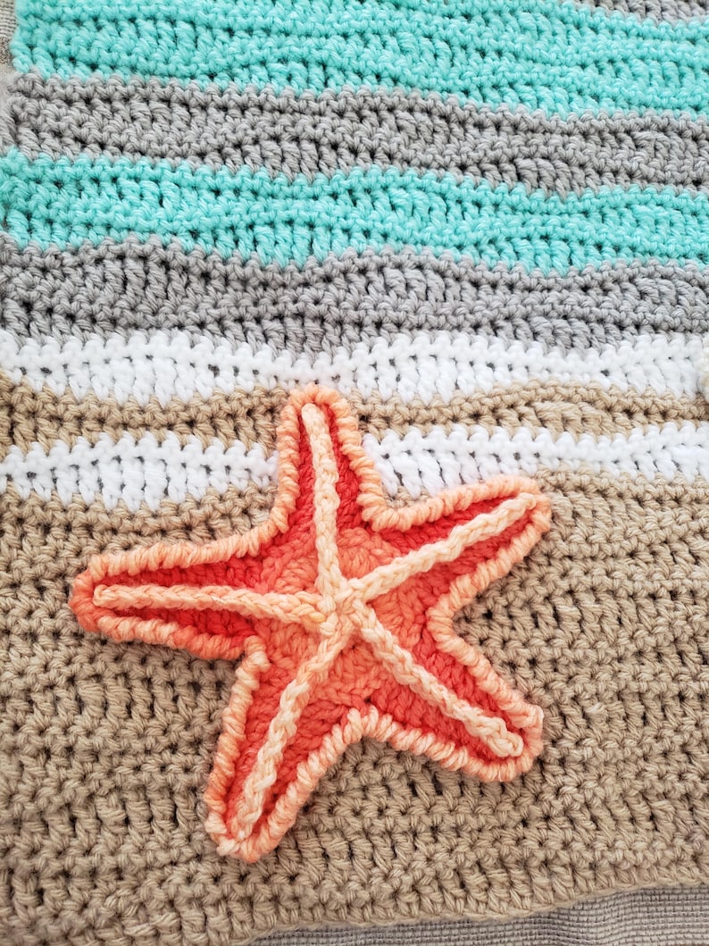 Peut inclure: Une couverture au crochet avec un motif ray&eacute; dans les tons de gris, blanc et turquoise. Une grande &eacute;toile de mer orange est appliqu&eacute;e sur la couverture.