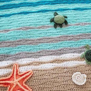May include: Crocheted beach scene blanket featuring a starfish, seashells, and turtles. The blanket is made with blue, green, white, and brown yarn.