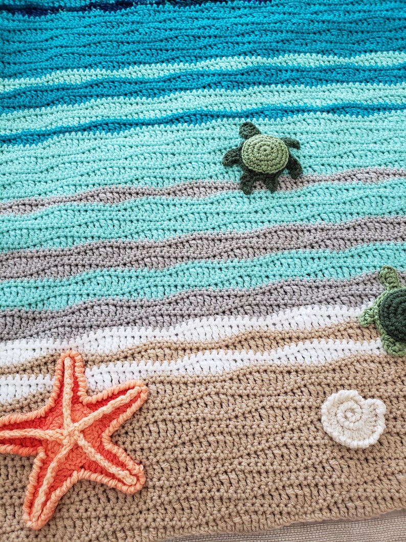 Peut inclure: Couverture en crochet repr&eacute;sentant une sc&egrave;ne de plage avec une &eacute;toile de mer, des coquillages et des tortues. La couverture est r&eacute;alis&eacute;e avec du fil bleu, vert, blanc et marron.