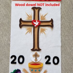 May include: A white felt banner with the name "ERIK" in black felt letters and the year "2020" in black felt numbers. The banner also features a brown felt cross with a white felt dove inside a red felt heart, a white felt communion wafer with a brown felt cross, a gold felt chalice with red felt wine, a purple felt bunch of grapes, and two brown felt wheat stalks. The text "Wood dowel NOT included" is printed in red felt letters at the bottom of the banner.