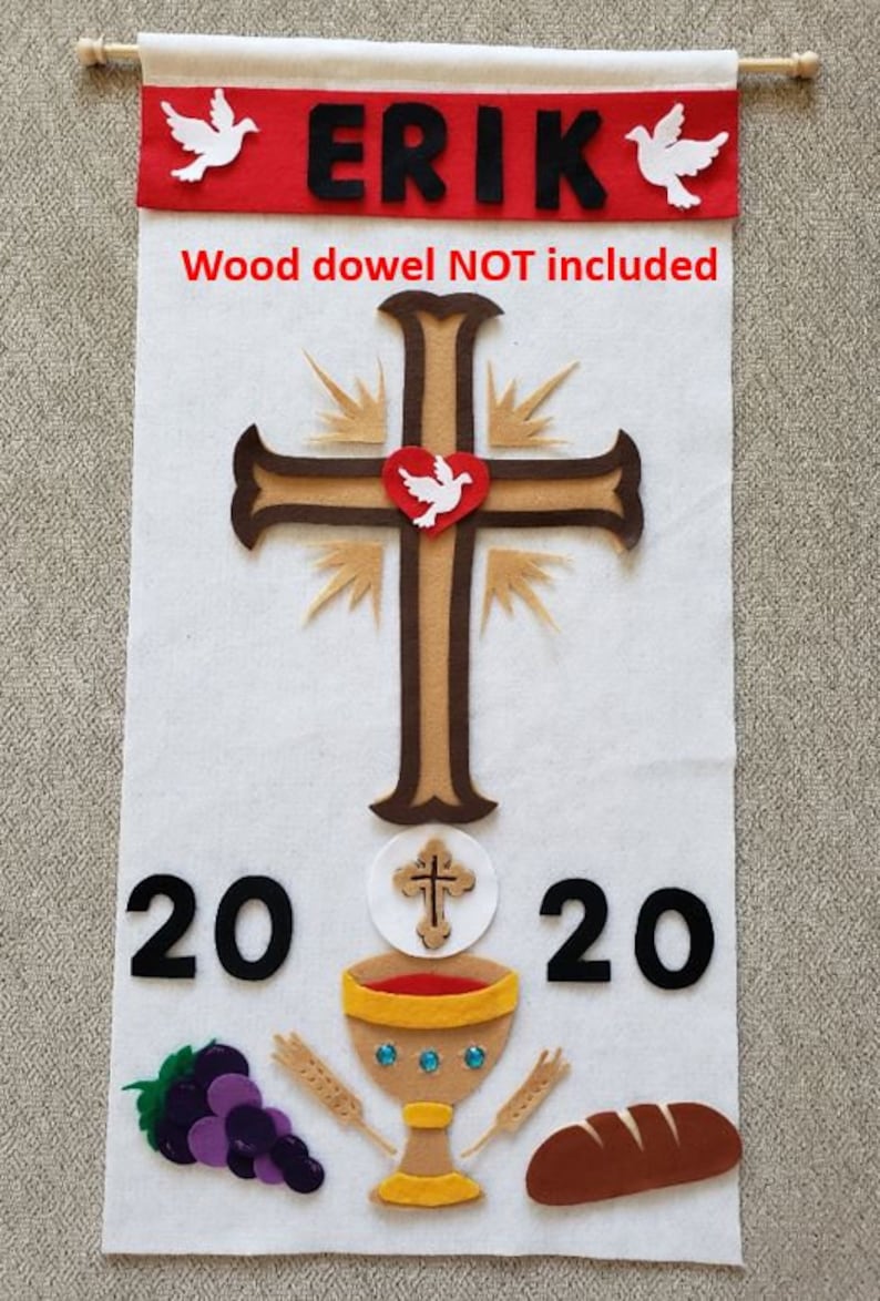 May include: A white felt banner with the name "ERIK" in black felt letters and the year "2020" in black felt numbers. The banner also features a brown felt cross with a white felt dove inside a red felt heart, a white felt communion wafer with a brown felt cross, a gold felt chalice with red felt wine, a purple felt bunch of grapes, and two brown felt wheat stalks. The text "Wood dowel NOT included" is printed in red felt letters at the bottom of the banner.