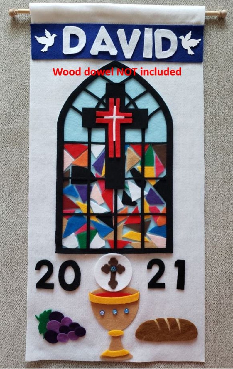 May include: A white felt banner with the name "DAVID" in blue felt letters and two white doves. A stained glass window design with a red cross is in the center. The banner also includes the year "2021", a chalice, a loaf of bread, and a bunch of grapes.