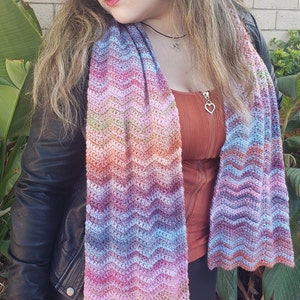 May include: A multi-colored crocheted scarf with a wavy pattern. The scarf is made with a variety of shades of pink, blue, green, and orange yarn.