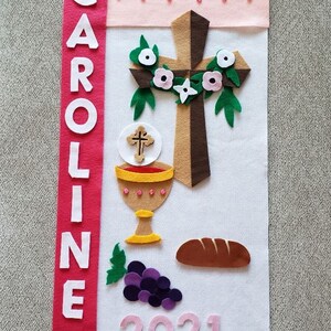 May include: White felt banner with the name CAROLINE in white letters on a red background. The banner also features a brown felt cross with a white felt flower wreath, a gold chalice with a red wine inside, a brown felt loaf of bread, a bunch of purple grapes, and the year 2021 in pink felt. The banner has a pink and red scalloped border. Wood dowel not included.
