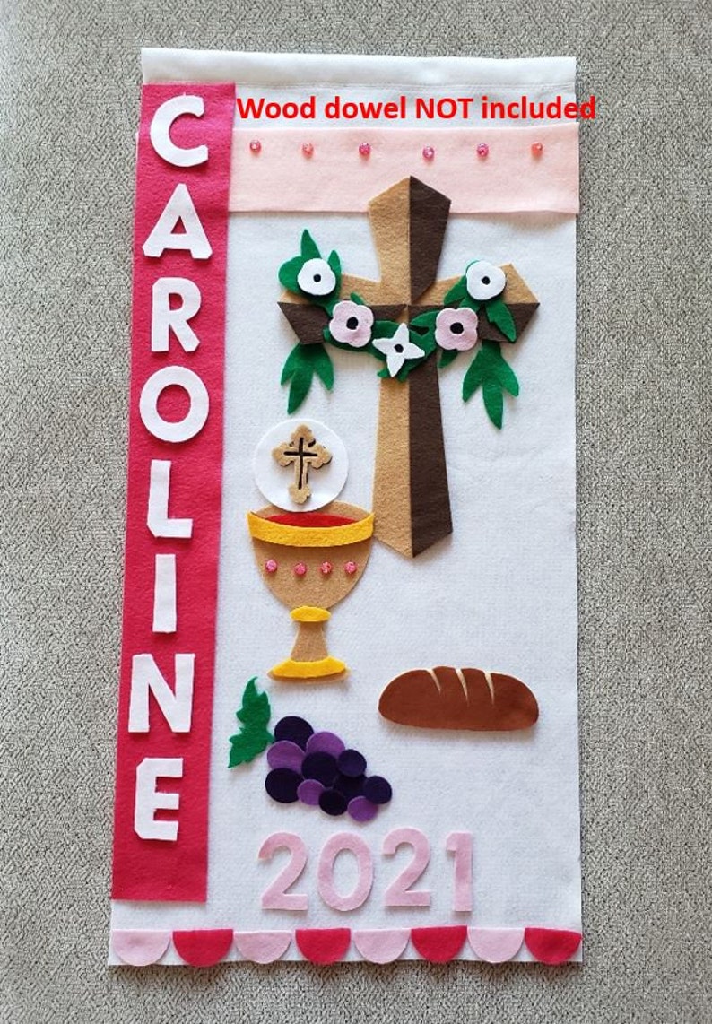 May include: White felt banner with the name CAROLINE in white letters on a red background. The banner also features a brown felt cross with a white felt flower wreath, a gold chalice with a red wine inside, a brown felt loaf of bread, a bunch of purple grapes, and the year 2021 in pink felt. The banner has a pink and red scalloped border. Wood dowel not included.