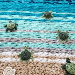 Peut inclure: Une couverture au crochet sur le th&egrave;me de la plage. La couverture pr&eacute;sente un oc&eacute;an bleu et vert avec des vagues blanches et un fond de sable brun. Il y a cinq tortues de mer au crochet vertes sur la couverture.