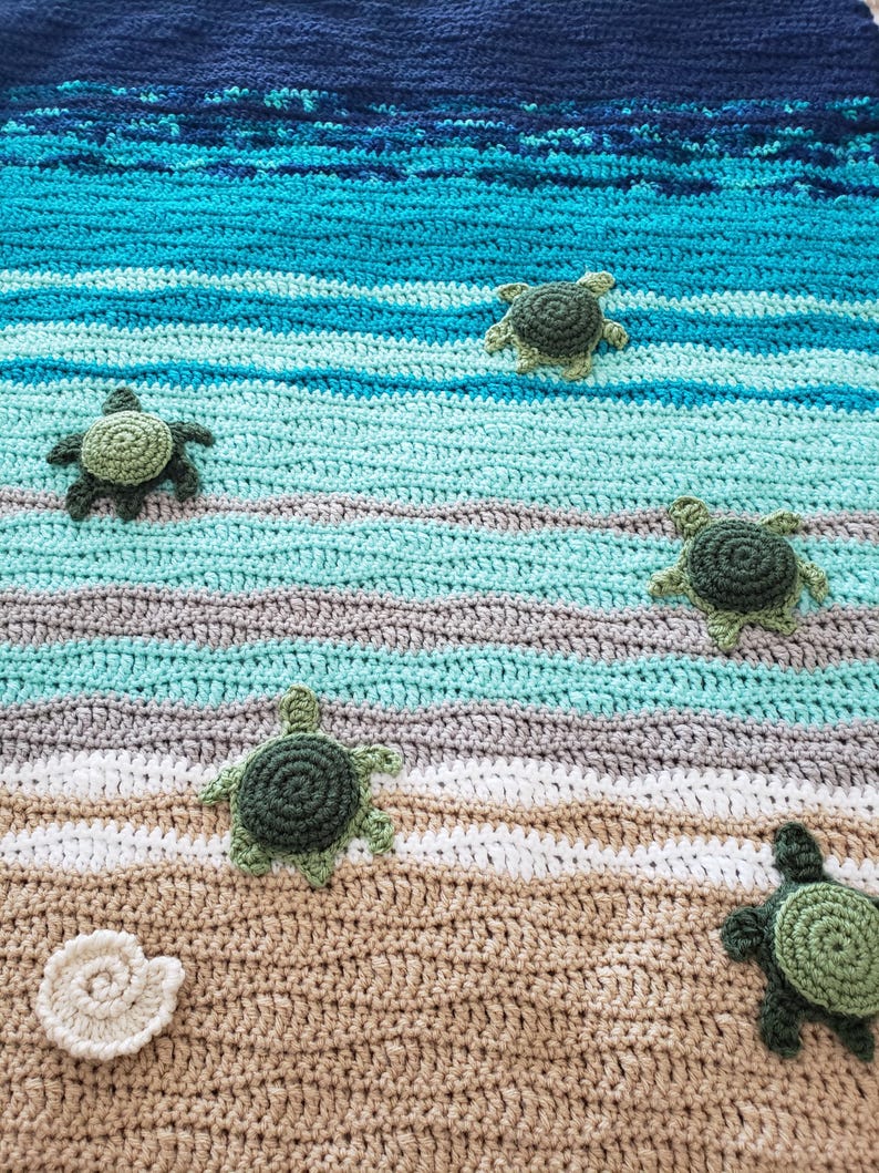 Peut inclure: Une couverture au crochet sur le th&egrave;me de la plage. La couverture pr&eacute;sente un oc&eacute;an bleu et vert avec des vagues blanches et un fond de sable brun. Il y a cinq tortues de mer au crochet vertes sur la couverture.