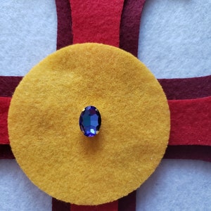 Pu&ograve; includere: Primo piano di un gioiello blu incastonato in oro, al centro di un cerchio di feltro giallo. Il cerchio &egrave; circondato da feltro rosso e bordeaux.