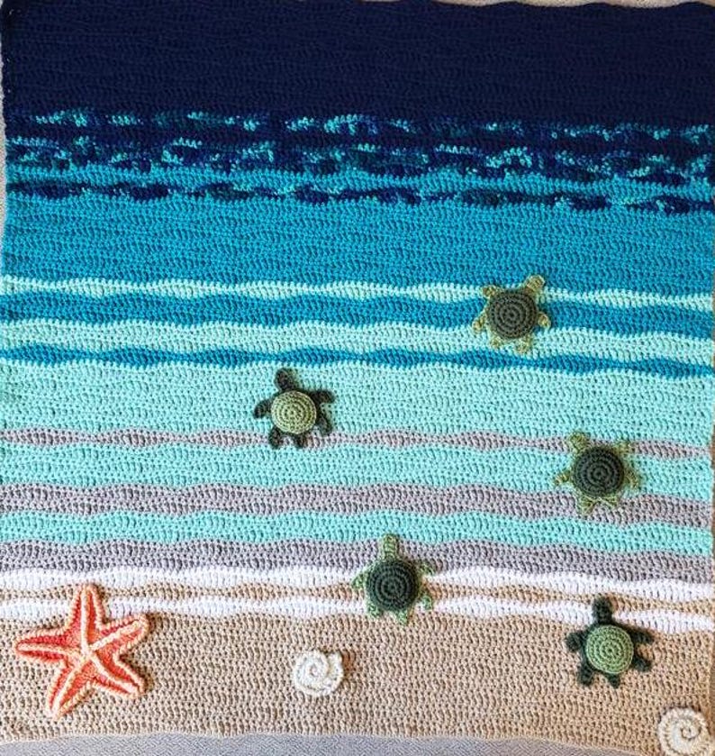 Peut inclure: Une couverture au crochet repr&eacute;sentant une sc&egrave;ne de plage. La couverture pr&eacute;sente une mer bleue et verte avec des vagues blanches, une plage de sable beige, une &eacute;toile de mer et cinq tortues de mer vertes.