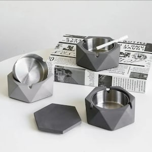 Peut inclure: Trois cendriers hexagonaux en béton gris avec un bol intérieur en acier inoxydable. Un cendrier a une cigarette posée sur le bord.