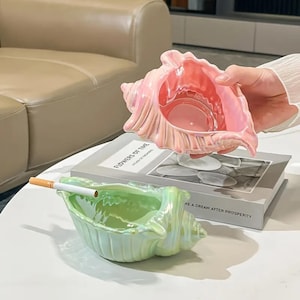 May include: Two seashell-shaped ashtrays, one pink and one green, sit on a table. The pink ashtray is held up by a person. A lit cigarette rests in the green ashtray. The ashtrays are iridescent and have a glossy finish.