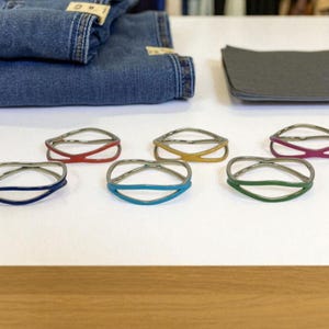 May include: Six colorful bracelets with a silver metal band and a colored, curved center. The bracelets are in shades of red, yellow, blue, teal, green, and purple. A pair of blue jeans and a gray cloth are in the background.