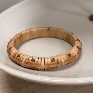 May include: A woven bamboo bracelet with a natural, light brown color. The bracelet is circular and rests on a white ceramic plate. The weave pattern is visible, showcasing the texture and craftsmanship of the accessory.