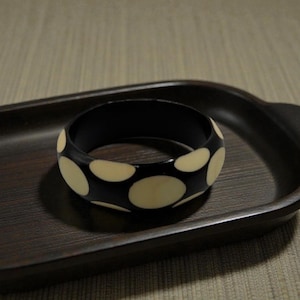 May include: A black and cream-colored bangle bracelet with a polka dot pattern. The bracelet is made of a glossy material and sits on a dark brown wooden tray.