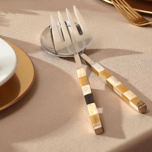 Può includere: Un set da portata per insalata in acciaio inossidabile con forchetta e cucchiaio. I manici presentano un motivo a mosaico di quadrati beige, neri e bianchi. Il set è esposto su una tovaglia beige, con un piatto bordato d'oro e un piatto bianco sullo sfondo.