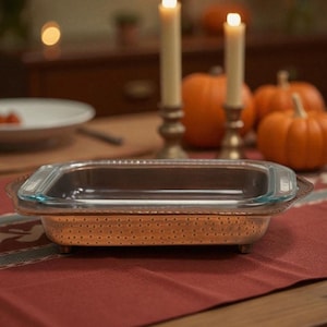 May include: A rectangular glass baking dish with a copper-colored metal base and a clear glass lid. The dish is on a red tablecloth, with pumpkins and lit candles in the background. A fall-themed kitchenware item.