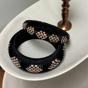 May include: Two black woven bracelets with a checkered pattern in natural tones rest on a white oval dish. The bracelets are thick and appear to be made of tightly wound threads. A small bronze candlestick is in the background.