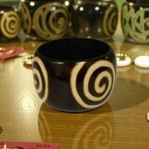 May include: A dark brown bangle bracelet with a cream-colored spiral design. Several other bracelets with similar designs are in the background. The bracelets are on a wooden surface, with jewelry and other accessories visible.