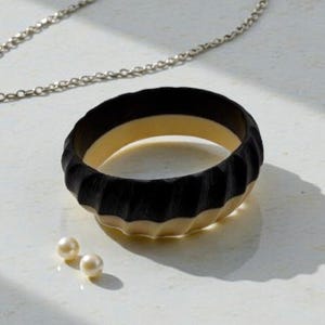 Puede incluir: Un brazalete negro y crema con un diseño acanalado. Dos pequeñas perlas redondas de color crema están al lado del brazalete. Una cadena plateada está en el fondo.
