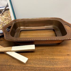 May include: A rectangular copper-coloured baking dish with handles and a decorative border. The dish sits on a wooden surface, with a wooden spoon and a cookbook visible in the background. The dish is empty.