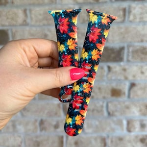 May include: Two colorful Apple Watch bands with a fall leaf pattern. The bands are black with red, orange, and yellow leaves on a dark background.