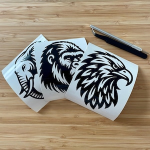May include: Three white vinyl stickers featuring black animal designs: an elephant head, a gorilla face, and an eagle head. A silver and black precision tool is also present. The stickers are on a wooden surface.