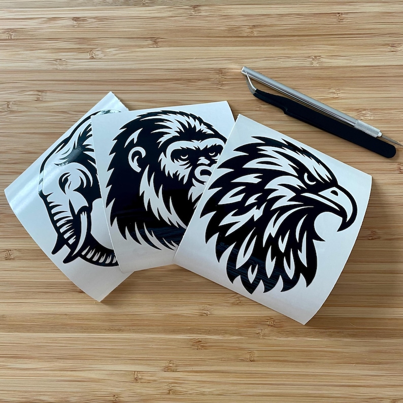 May include: Three white vinyl stickers featuring black animal designs: an elephant head, a gorilla face, and an eagle head. A silver and black precision tool is also present. The stickers are on a wooden surface.