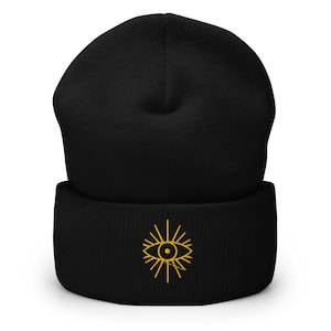 Puede incluir: Gorro negro con un diseño de ojo bordado en oro. El ojo tiene un patrón de rayos de sol alrededor.