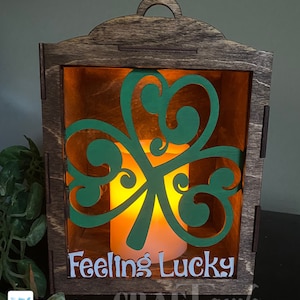 Pu&ograve; includere: Una lanterna di legno con un ritaglio a forma di trifoglio verde e il testo "Feeling Lucky" sul davanti. La lanterna &egrave; illuminata da una candela all'interno.